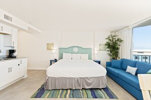 Habitación de hotel con cama, sofá azul, zona de cocina y un ventanal con cortinas.