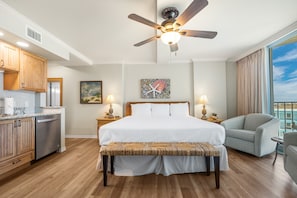 A hotel room with a bed, a bench, a ceiling fan, a view of the ocean, and a kitchenette. - Sandcastle Condominiums Unit 306 (Port Aransas)