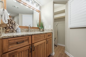A bathroom with wooden cabinetry, a granite countertop, a mirror with lights, a towel rack, and a plant. - Sandcastle Condominiums Unit 306 (Port Aransas)
