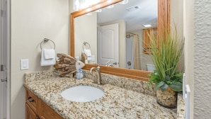 Une salle de bain avec un comptoir en granit, un lavabo blanc, un meuble-lavabo en bois, un grand miroir et une plante en pot.