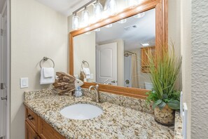 Un baño con encimera de granito, un lavamanos blanco, un mueble de tocador de madera, un espejo grande y una planta en maceta.