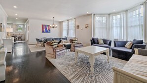 A spacious living room with dark wooden flooring, a white coffee table, and a sofa set. There is a dining area with a table and chairs, and a kitchen in the background.