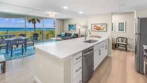 A modern kitchen with a large island, a dining area with a view of the beach, and a living area with a sofa and artwork.