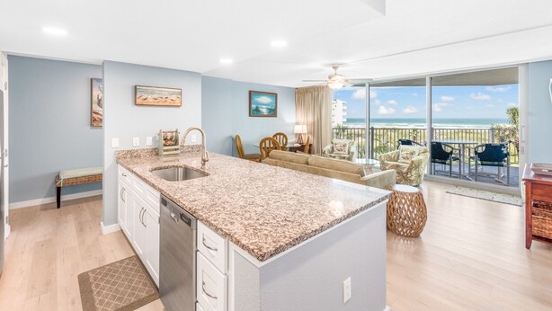 Una cocina moderna con encimeras de granito, lavavajillas integrado y una isla grande. Hay un comedor con vista al océano y un balcón con mobiliario de exterior.
