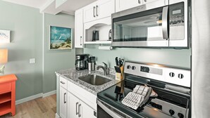 A modern kitchen with white cabinets, a granite countertop, stainless steel appliances, and a painting of a beach scene. - Sandcastle Condominiums Unit 328 (Port Aransas)