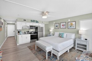 Un dormitorio con cama, mesitas de noche, escritorio, ventilador de techo y kitchenette.
