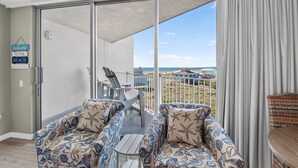 A balcony with a view of the beach, two chairs with a nautical pattern, a table, and a sign that reads 'Welcome to the Beach'.