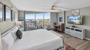 A hotel room with a large bed, a TV, a balcony with chairs, and a view of the beach.