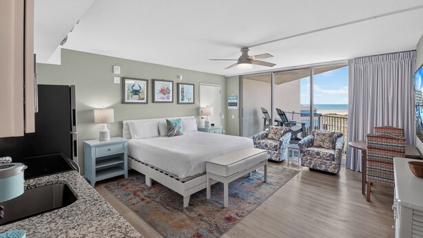 A hotel room with a bed, a desk, a chair, a balcony with a view of the beach, and a ceiling fan.