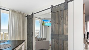 A room with a sliding barn door, a view of the outdoors, a television, and a dining table. - Sandcastle Condominiums Unit 337 (Port Aransas)