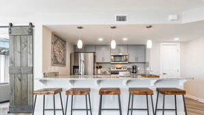 A modern kitchen with a central island, bar stools, stainless steel appliances, and pendant lighting. - Sandcastle Condominiums Unit 337 (Port Aransas)