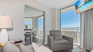 A modern hotel room with a balcony, a grey sofa, a small table with a lamp, and a TV displaying a pool view. - Sandcastle Condominiums Unit 337 (Port Aransas)