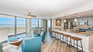 A spacious living area with a large window overlooking the ocean, a kitchen with modern appliances, and a bar area with stools. - Sandcastle Condominiums Unit 337 (Port Aransas)