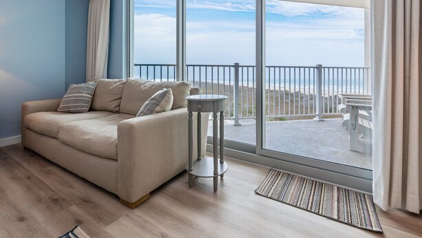 A modern living room with a beige sofa, a small round table, and a view of a balcony with outdoor furniture. - Sandcastle Condominiums Unit 402 (Port Aransas)