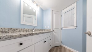 A bathroom with a granite countertop, white cabinets, a mirror with lights, and a door with blinds. - Sandcastle Condominiums Unit 402 (Port Aransas)