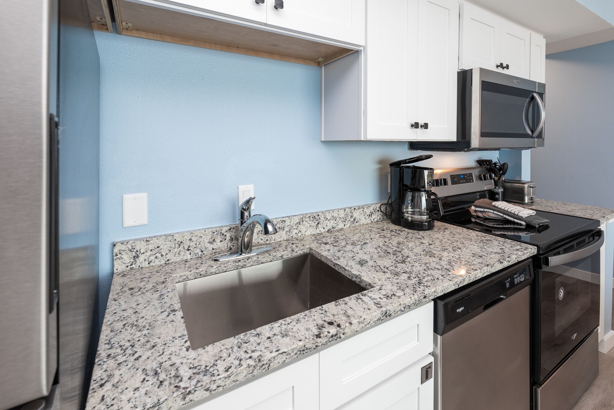 A kitchen with a granite countertop, stainless steel sink, white cabinets, a microwave, a coffee maker, and a dishwasher.