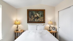 Un dormitorio con una cama grande, dos mesitas de noche con lámparas, un cuadro en la pared y una puerta a la derecha.