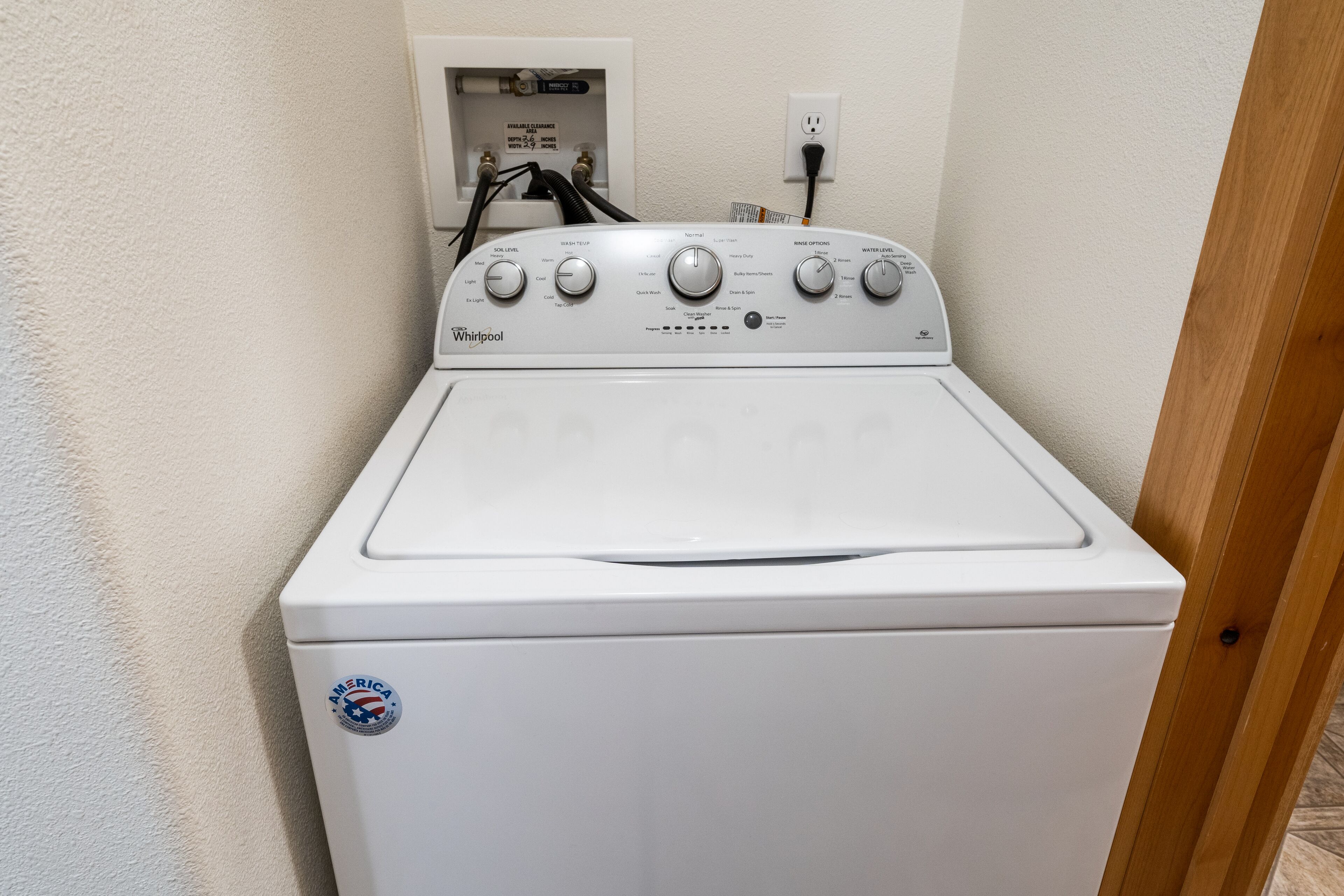 A front view of a Whirlpool washer with control knobs, a water inlet, and a power outlet on the wall.