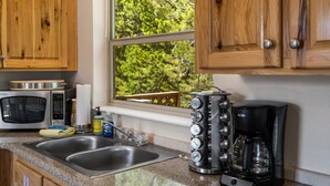 Una cocina con armarios de madera, un lavamanos de acero inoxidable, un microondas, una cafetera y una ventana con vista a árboles.