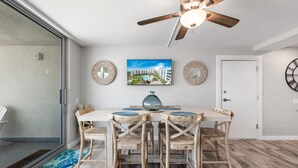 A dining area with a wooden table and chairs, a ceiling fan, and a large window. - Sandcastle Condominiums Unit 404 (Port Aransas)