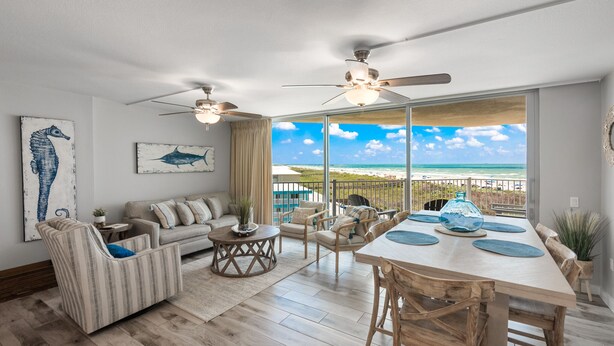 A spacious living room with a large window overlooking a beach, a ceiling fan, and a dining area with a wooden table and chairs. - Sandcastle Condominiums Unit 404 (Port Aransas)