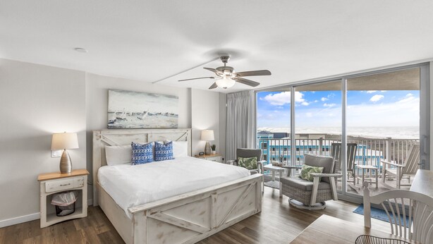 A bedroom with a bed, a ceiling fan, a nightstand, a chair, and a view of the ocean. - Sandcastle Condominiums Unit 410 (Port Aransas)