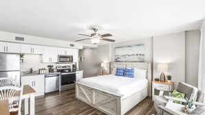 A modern hotel room with a kitchenette, a bed with white bedding, a ceiling fan, and a painting on the wall. - Sandcastle Condominiums Unit 410 (Port Aransas)