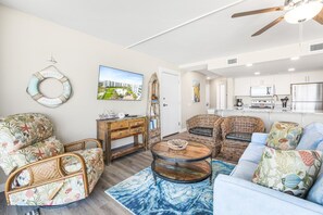 A living room with a sofa, armchair, and a coffee table. There is a television mounted on the wall and a kitchen area visible through an open door. - Sandcastle Condominiums Unit 412 (Port Aransas)
