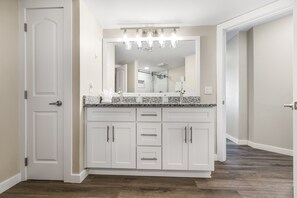 A modern bathroom with a large vanity, a mirror with lights, and a shower area visible in the background. - Sandcastle Condominiums Unit 412 (Port Aransas)