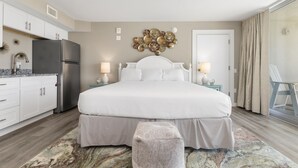 A bedroom with a large bed, a ceiling fan, a refrigerator, a nightstand with lamps, and a view of the outside. - Sandcastle Condominiums Unit 415 (Port Aransas)