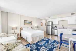 A bedroom with a bed, bedside tables, a desk, and a chair. There is a kitchen area with a refrigerator, stove, and sink. - Sandcastle Condominiums Unit 419 (Port Aransas)