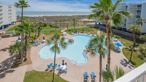 A resort pool area with palm trees, lounge chairs, and a view of the beach. - Sandcastle Condominiums Unit 420 (Port Aransas)