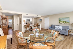 A spacious living room with a glass-top dining table surrounded by wicker chairs, a blue sofa, and a painting of a beach scene. - Sandcastle Condominiums Unit 420 (Port Aransas)