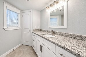 A bathroom with a white door, a vanity with a granite countertop, a mirror, and a window with blinds. - Sandcastle Condominiums Unit 428 (Port Aransas)