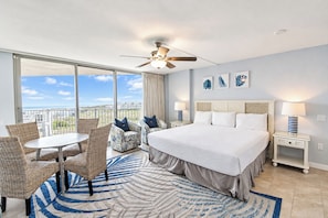 Quarto de hotel com uma cama grande, varanda com vista, ventilador de teto e área de jantar com móveis de vime.