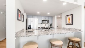 A modern kitchen with a granite countertop, stainless steel appliances, and wooden stools. - Sandcastle Condominiums Unit 430 (Port Aransas)