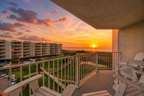 A balcony with a view of a sunset over the ocean, a pool area, and a building with multiple floors.
