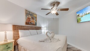 A bedroom with a bed, a ceiling fan, a painting, and a TV displaying a beach scene.