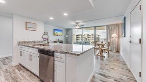 A modern kitchen with a dishwasher, white cabinets, and a granite countertop. There is a dining area with a table and chairs, and a balcony with a view of buildings.