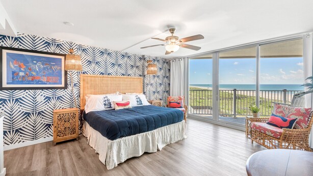 A bedroom with a large bed, a ceiling fan, a window with a view of the ocean, and a wicker chair with a cushion. - Sandcastle Condominiums Unit 502 (Port Aransas)
