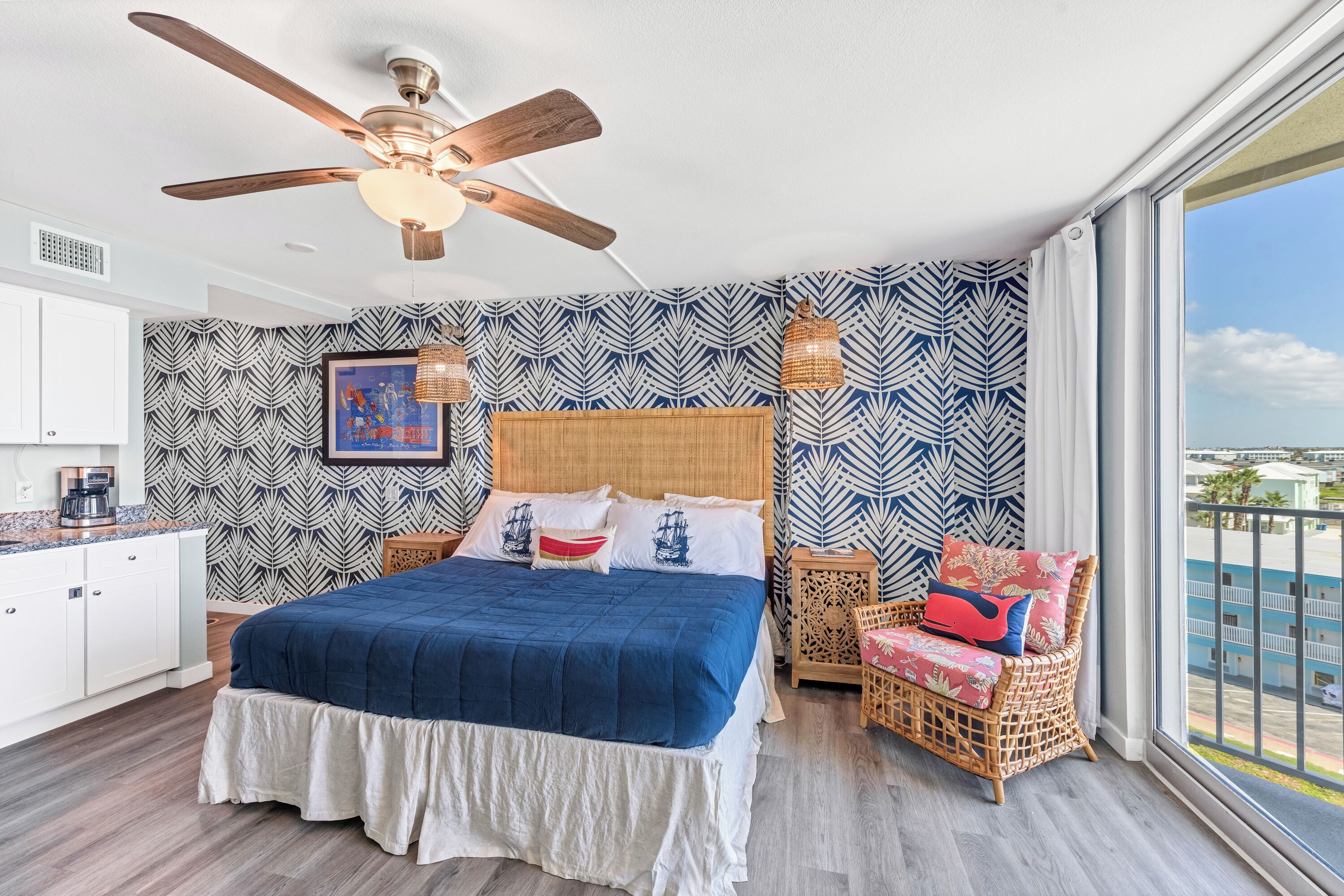 A bedroom with a bed, a ceiling fan, a window with a view, and patterned wallpaper.