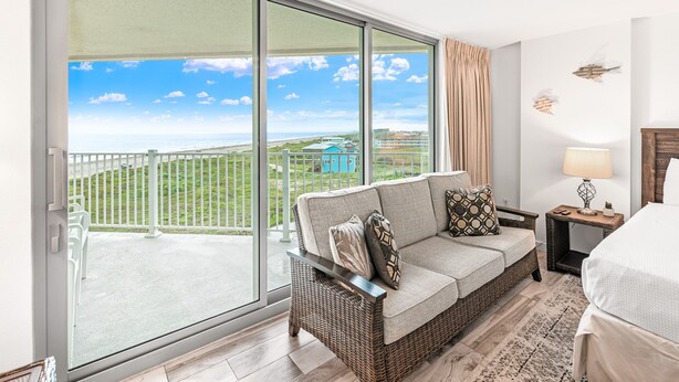 A balcony with a view of a green landscape and buildings, a wicker sofa with cushions, a small table with a lamp, and a bed with a white cover. - Sandcastle Condominiums Unit 503 (Port Aransas)