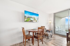 A dining area with a round wooden table and four chairs, a painting of a resort on the wall, and a balcony with white chairs. - Sandcastle Condominiums Unit 503 (Port Aransas)