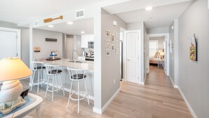 Une cuisine moderne avec des tabourets de bar blancs, un coin repas avec une lampe, et un couloir menant à une chambre.