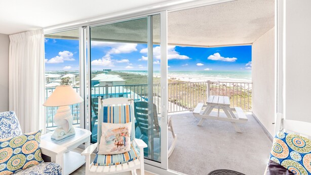 Un balcon donnant sur la plage, une chaise avec un coussin, une petite table avec une lampe et une fenêtre avec des rideaux.