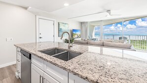 A modern kitchen with granite countertops, a double sink, and white cabinetry. There is a sliding glass door leading to a balcony with a view of the ocean. - Sandcastle Condominiums Unit 509 (Port Aransas)
