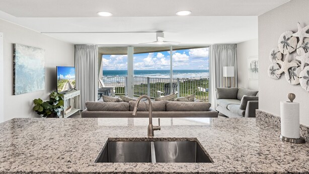 A modern kitchen with a large window overlooking the beach, a built-in sink, and a seating area with a view. - Sandcastle Condominiums Unit 509 (Port Aransas)