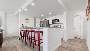 A modern kitchen with white cabinetry, a central island with red bar stools, stainless steel appliances, and a wooden floor. - Sandcastle Condominiums Unit 518 (Port Aransas)