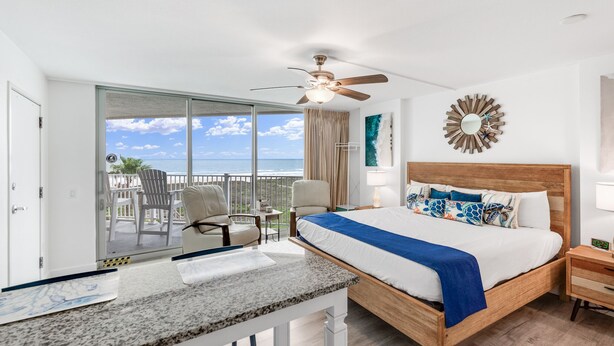 A hotel room with a bed, a desk, a chair, a balcony with a view of the ocean, and a ceiling fan. - Sandcastle Condominiums Unit 519 (Port Aransas)