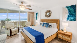 A bedroom with a large bed, a ceiling fan, a nightstand, a chair, and a view of the ocean. - Sandcastle Condominiums Unit 519 (Port Aransas)